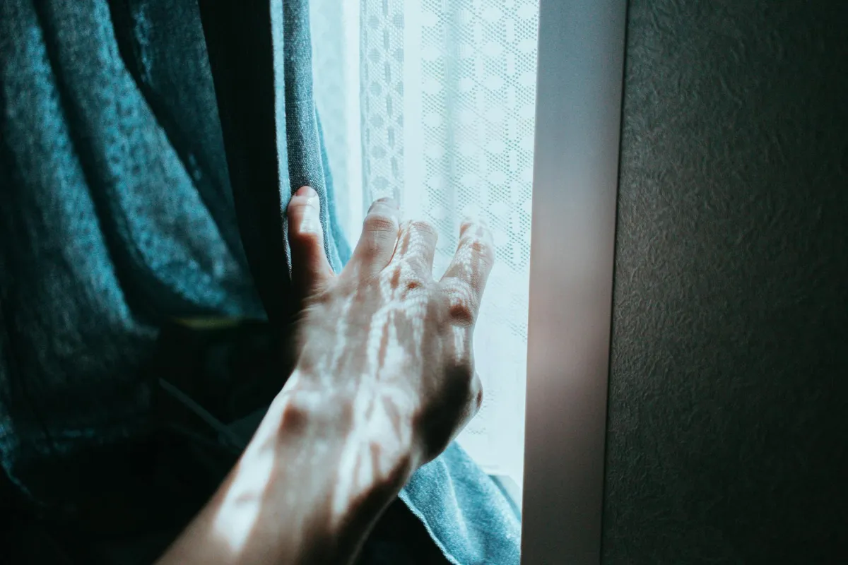 A hand gently opens a curtain, revealing sunlight streaming through a window indoors.